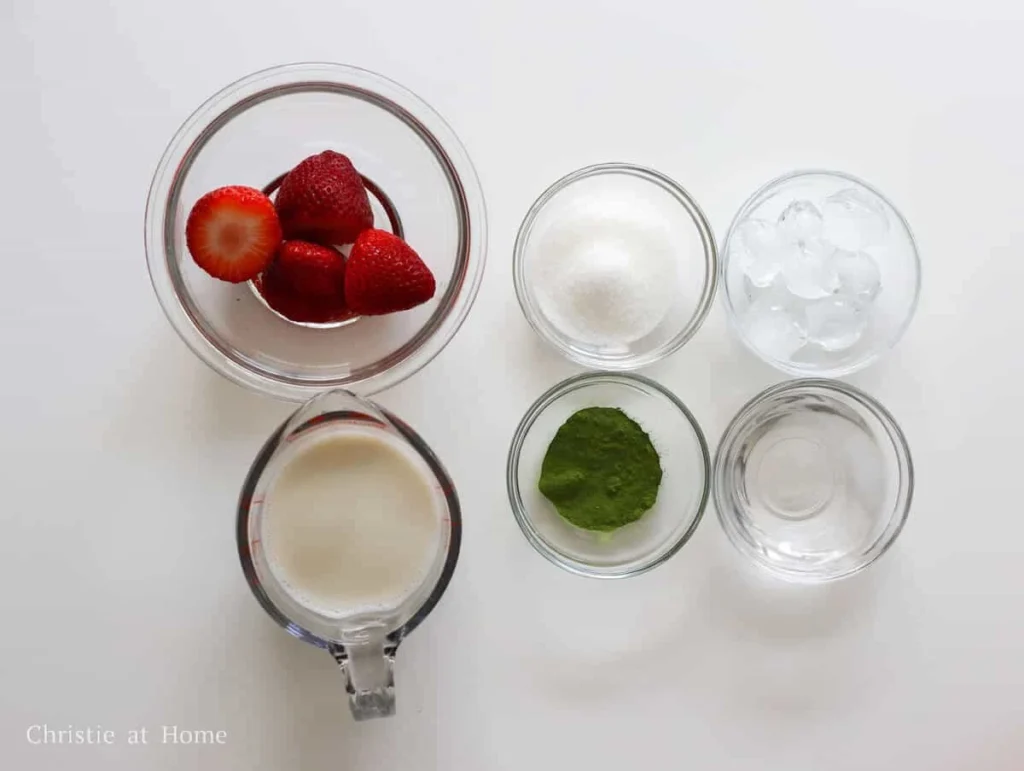 Ingredients for matcha strawberry latte including fresh strawberries, matcha powder, plant-based milk, and sugar laid out on a table